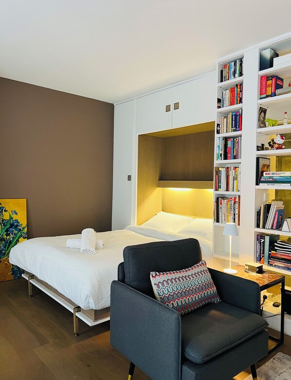 Relax in a serene space with a neatly made bed, comfortable armchair, and a well-organized bookshelf.