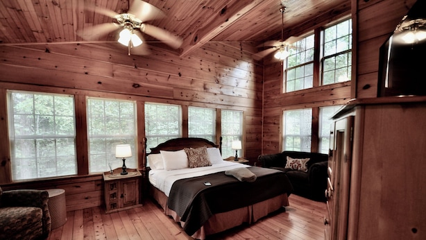 A wooden cabin bedroom with a bed, nightstands, a chair, and large windows. - Trails End, your own private retreat in the Smokies! (Townsend)