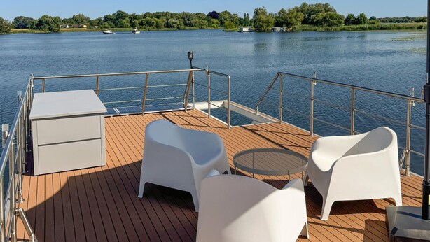 Une terrasse avec une table et des chaises, un petit meuble et une rambarde donnant sur une étendue d’eau où l’on aperçoit des bateaux et, au loin, de la végétation.