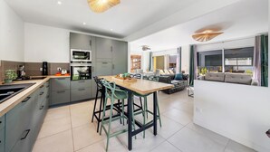 A modern kitchen with built-in appliances, a dining area with a wooden table and chairs, and a living room with a sofa and a large window.