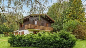 A two-story wooden house with a balcony, surrounded by lush greenery and trees.