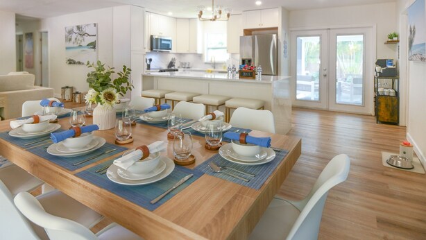 A dining area with a wooden table set for six, white chairs, and a kitchen in the background.