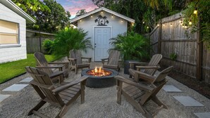A backyard with a fire pit surrounded by Adirondack chairs, a wooden shed with palm tree decorations, and a wooden fence.