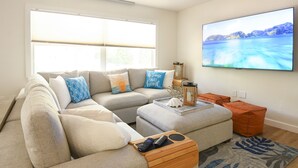 A living room with a sectional sofa, a coffee table, a TV mounted on the wall, and a large window with blinds.