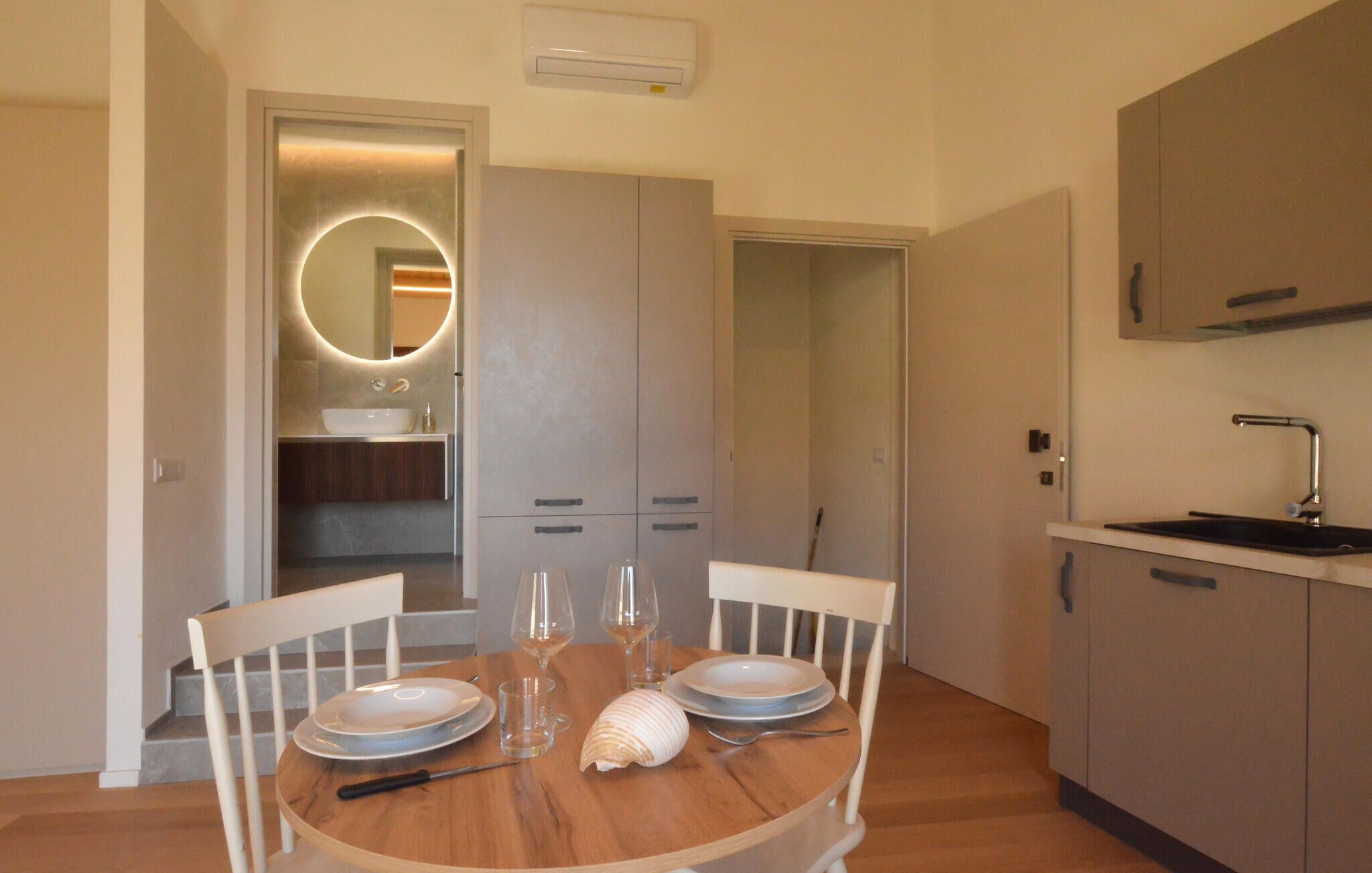 Un comedor con una mesa redonda, sillas blancas y piso de madera. Se observa una cocina con pileta y alacenas al fondo.