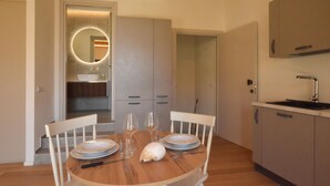 Un comedor con una mesa redonda, sillas blancas y piso de madera. Se observa una cocina con pileta y alacenas al fondo.