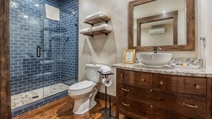 A bathroom with a glass shower enclosure, a white toilet, a marble sink, and wooden cabinets. - Bluebird Place Jessie Room: Historic Rt 66 (Elk City)