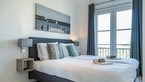 Una habitación de hotel con una cama grande, un cuadro de un lago, una ventana con cortinas y una mesita de noche con una lámpara.