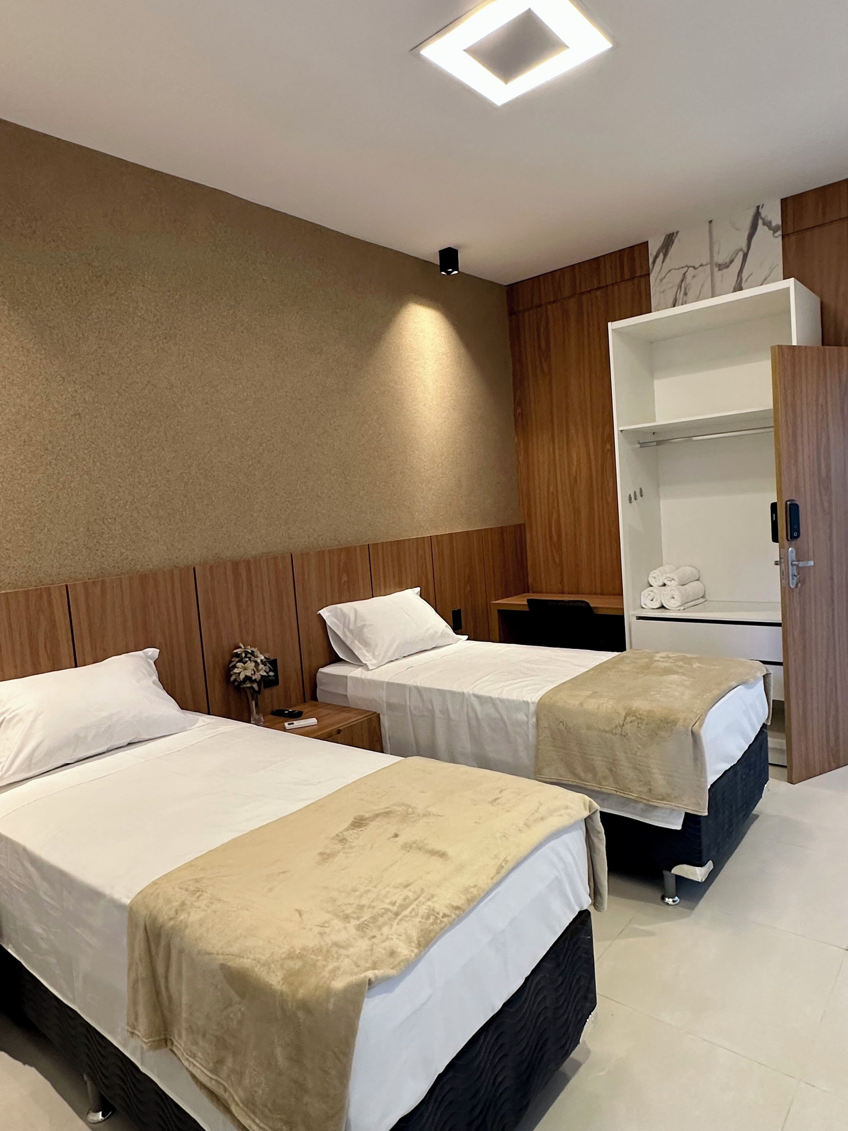 Habitación de hotel con dos camas, cabecero de madera, mesita de noche con lámpara, armario y luz en el techo.