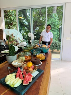 A buffet setup with various food items, a man standing behind it, and a view of greenery through large windows. - Pousada Alphavile (Palmas)