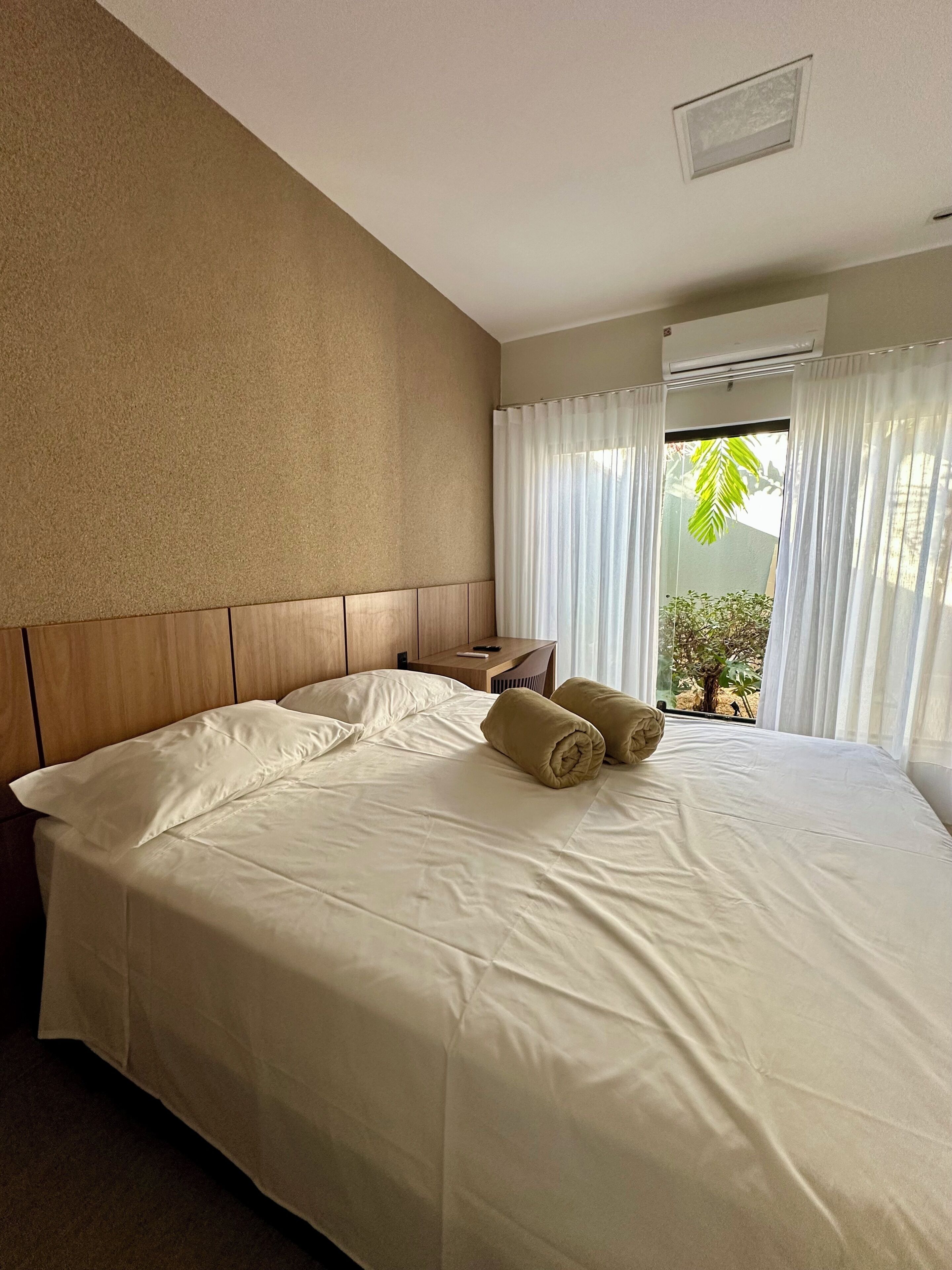 Una habitación de hotel con una cama grande, un cabecero de madera, una mesita de noche y un ventanal con cortinas.