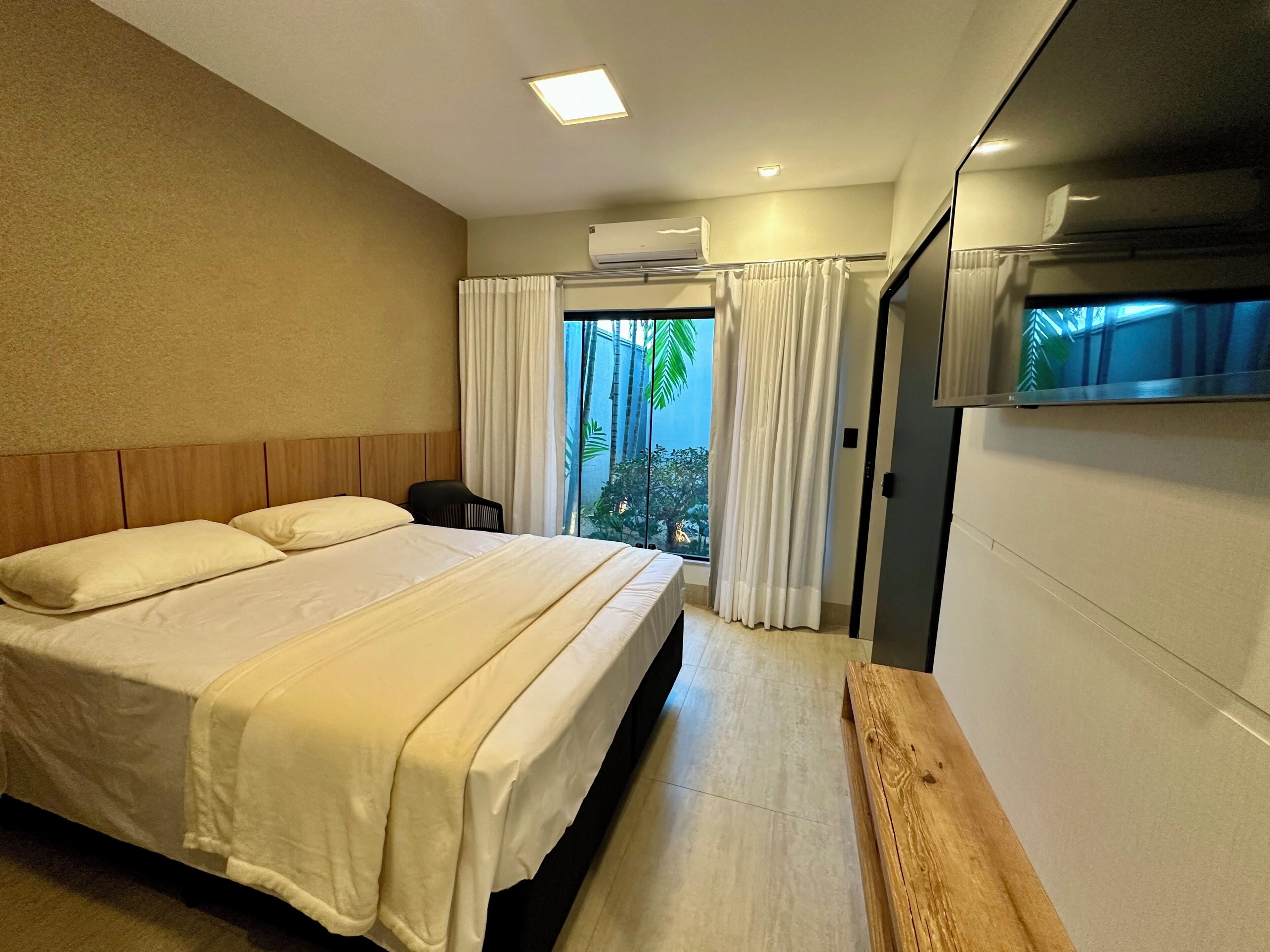 A hotel room with a bed, a small table, a window with curtains, and an air conditioning unit.