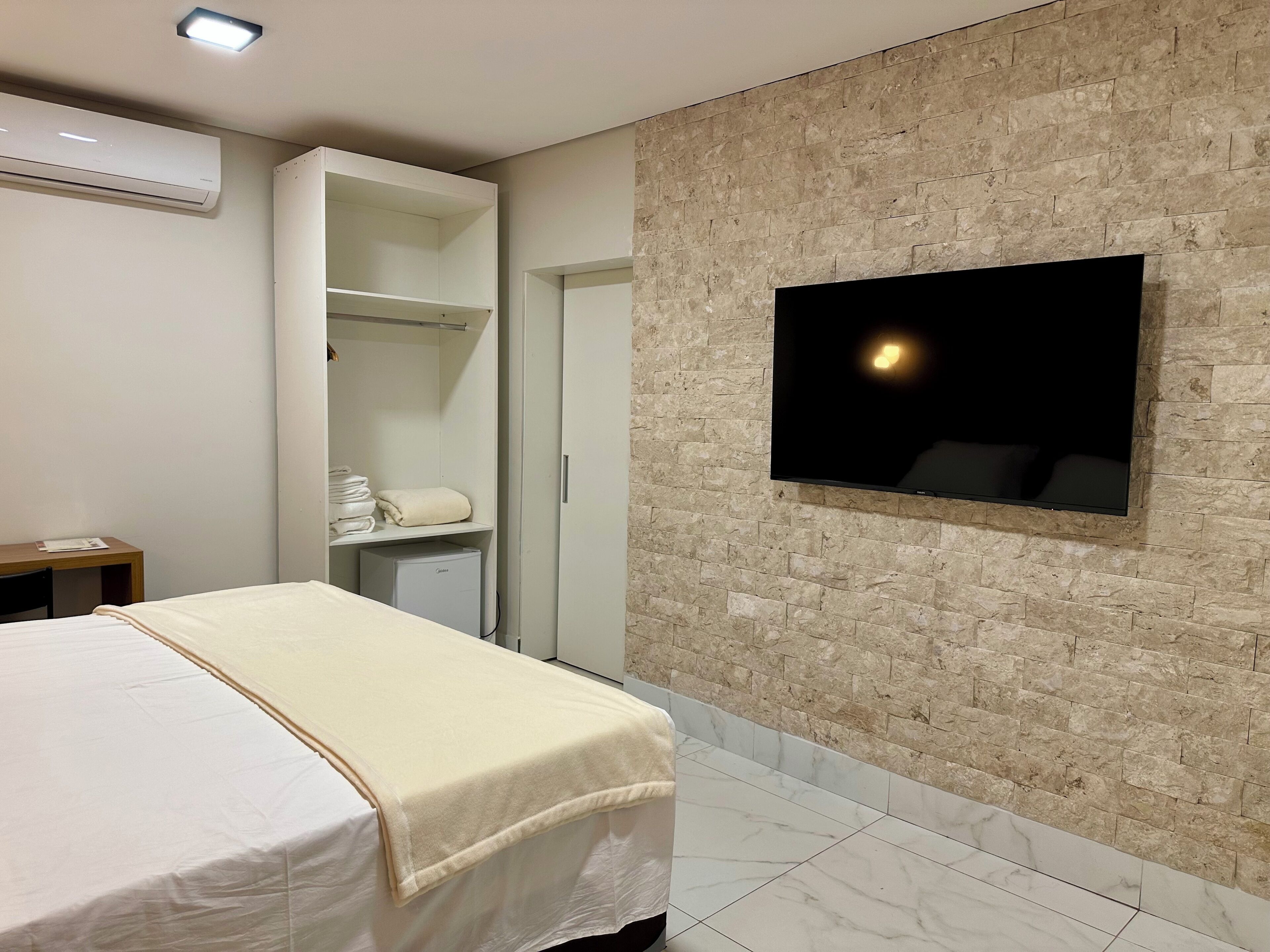 A hotel room with a bed, a TV mounted on a brick wall, a wardrobe, and a small desk.