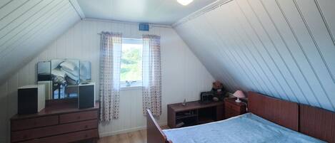 Een kleine slaapkamer, type zolderkamer, met een schuine kap, een houten bed, een dressoir, een bureau met stoel, een raam met bloemen gordijnen en een lamp.