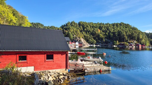 A red building with a black roof near a calm lake, surrounded by trees and docked boats. - Beautiful home in Reksteren with WiFi (Reksteren)
