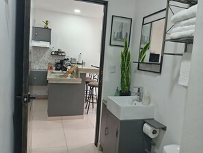 A modern kitchen with a central island, a sink, and a countertop. There is a dining area with chairs and a coffee machine. The bathroom includes a toilet, sink, and towel rack.