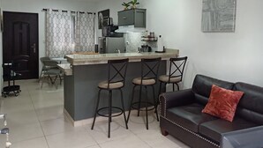 A modern living room with a black leather sofa, a kitchen island with bar stools, and a ceiling fan. - cr.arenalapartments #1 beautiful, comfortable with parking (La Fortuna)
