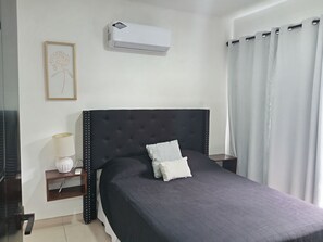 A bedroom with a dark upholstered headboard, a bedside table with a lamp, a framed picture on the wall, and a window with curtains.