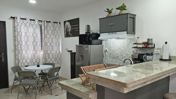 A modern kitchen with a central island, a dining area with chairs, a refrigerator, a microwave, and a small cabinet with plants. - cr.arenalapartments #1 beautiful, comfortable with parking (La Fortuna)