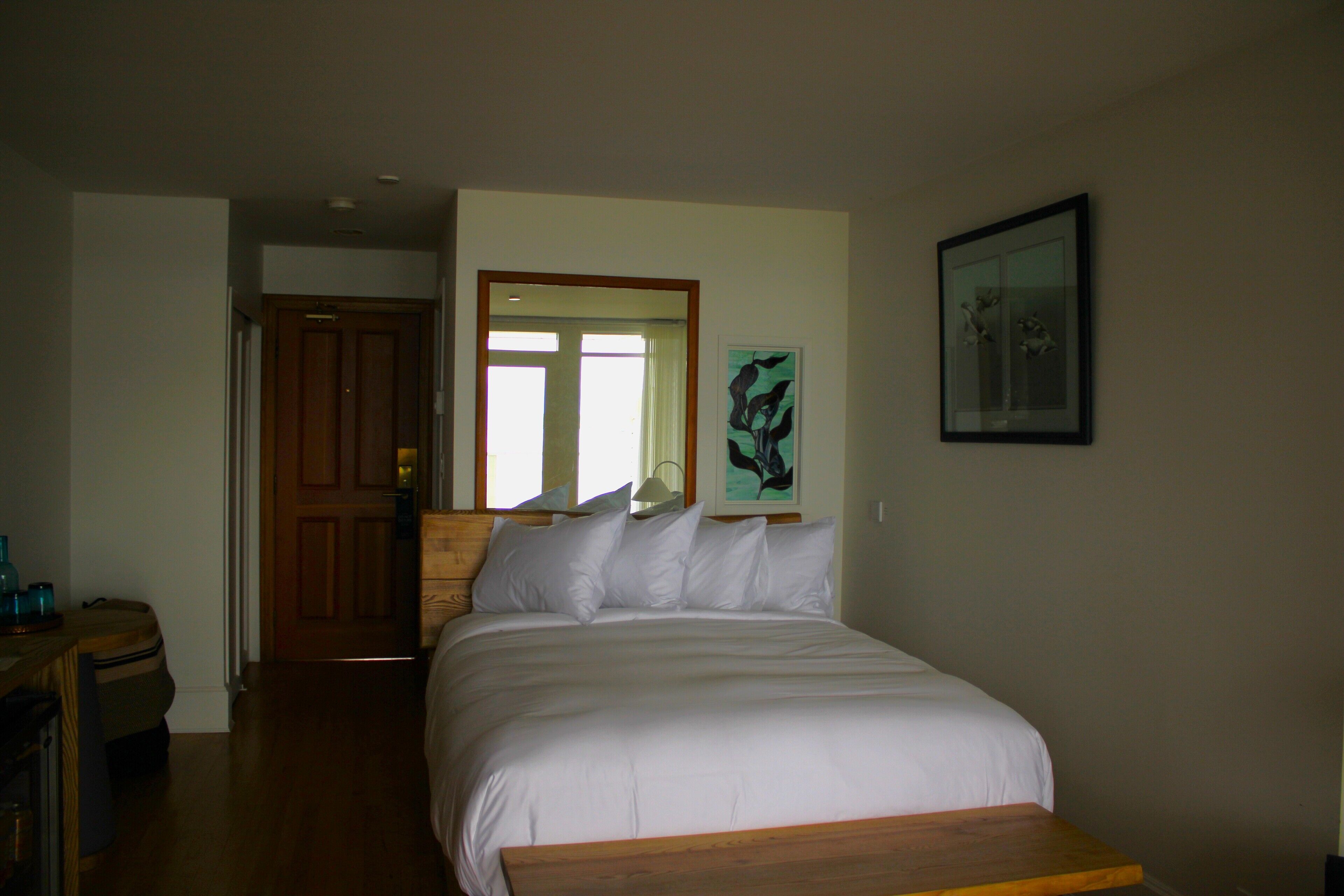 Quarto com cama de madeira, roupa de cama branca, mesa de cabeceira de madeira, quadro na parede e porta que dá para outro cômodo.
