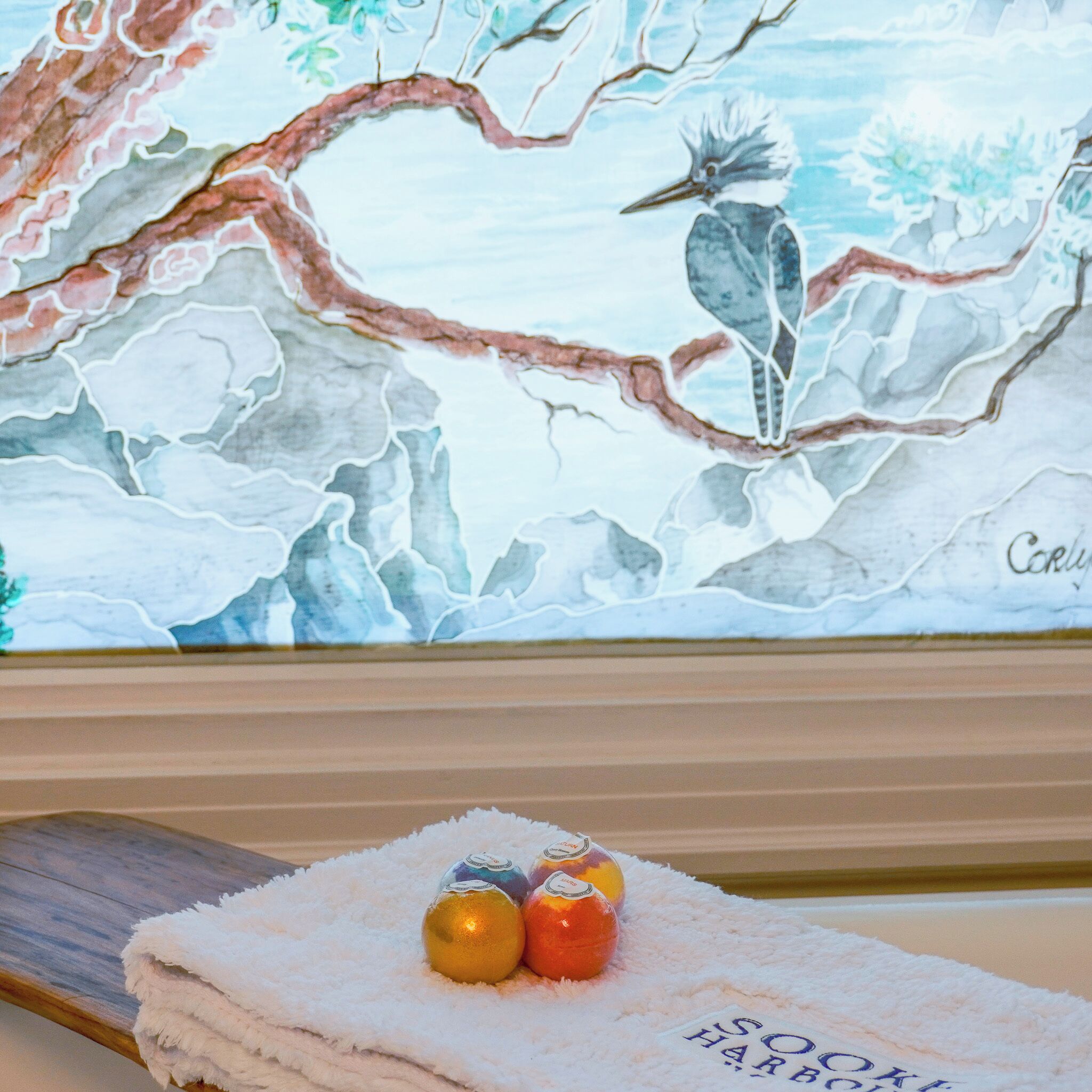 A towel with 'SOOK HOUSE' text, a painting of a bird on a tree, and two wrapped chocolates on the towel.