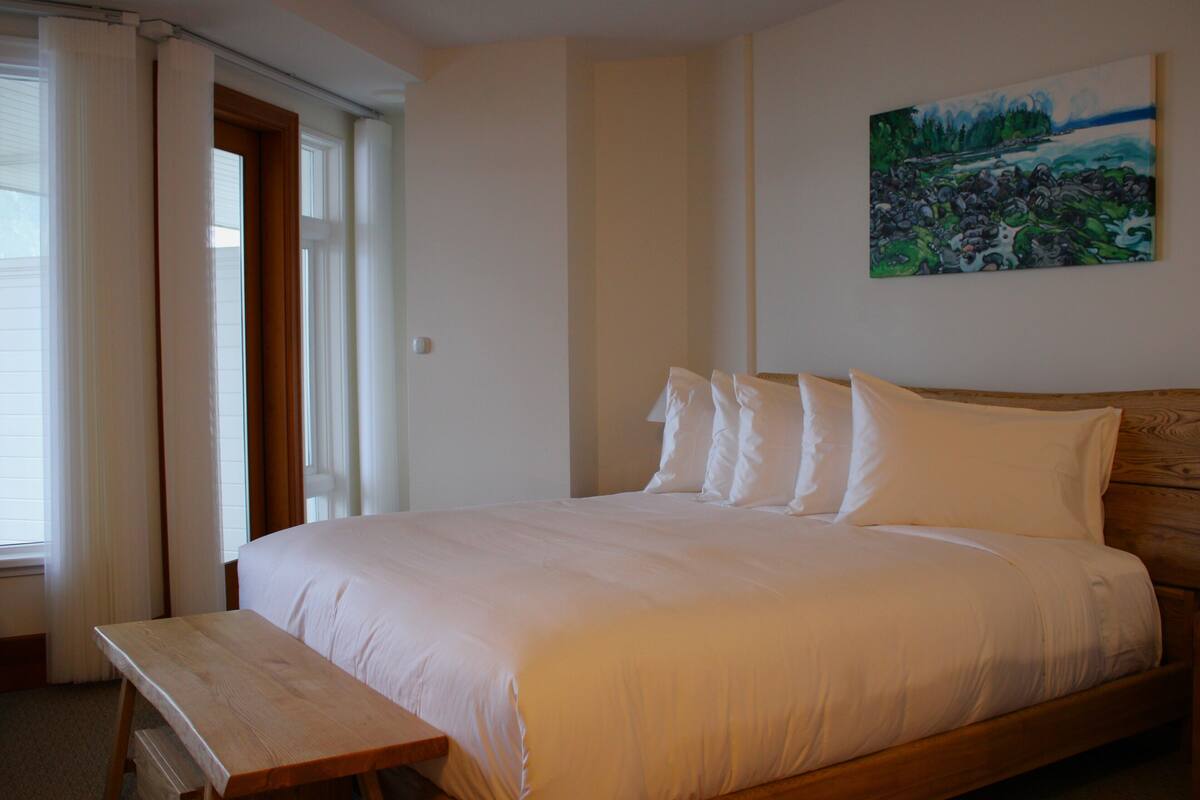 A bedroom with a wooden bed, white bedding, a bench at the foot of the bed, and a painting on the wall.