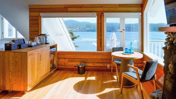 A cozy cabin with a wooden interior, a dining area with a table and chairs, and a view of a lake and mountains through large windows. - Sooke Harbour House (Sooke)