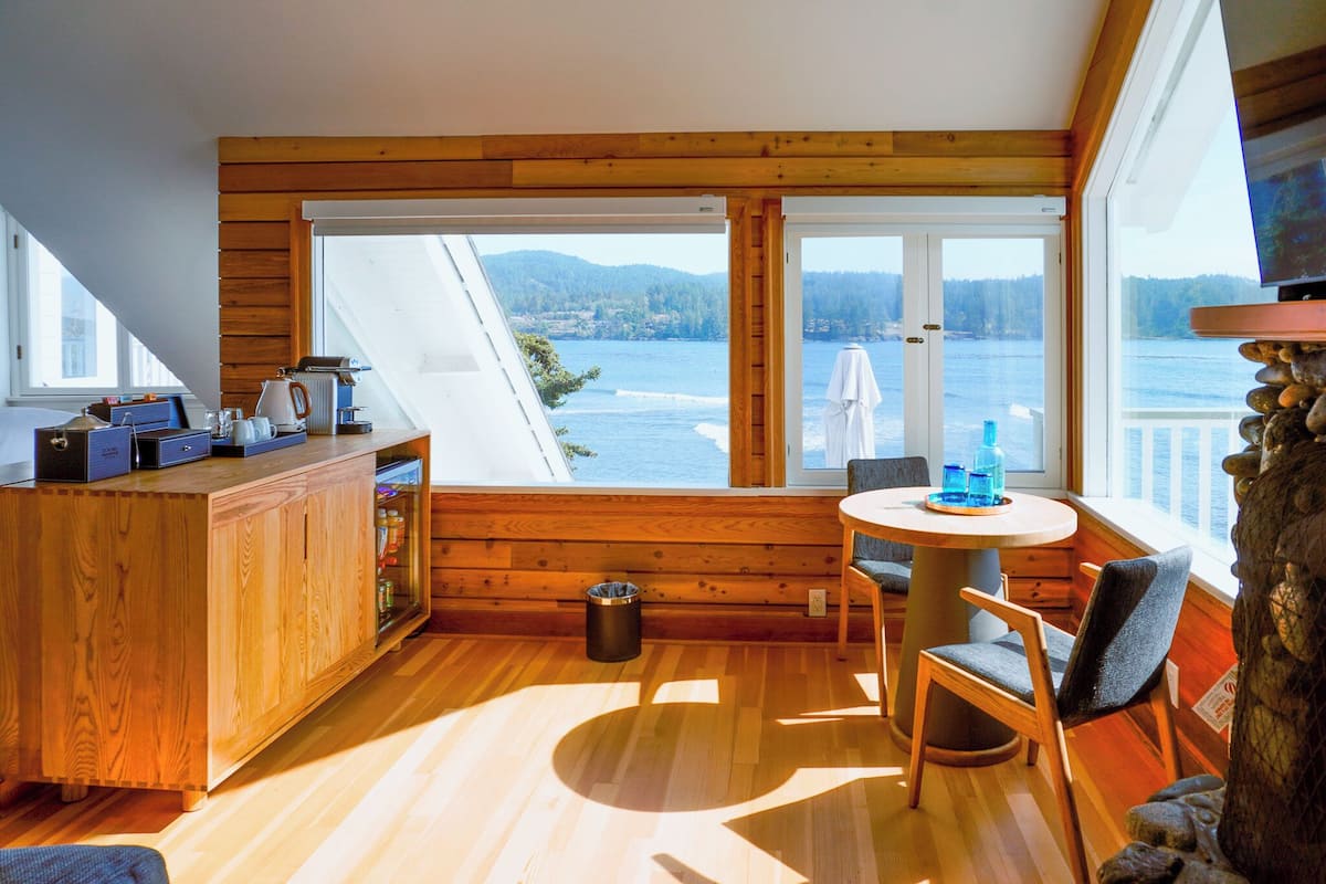 A cozy cabin with a wooden interior, a dining area with a table and chairs, and a view of a lake and mountains through large windows.