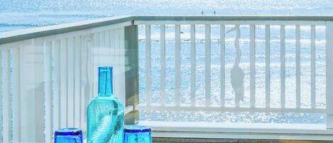 Un balcone con un tavolo, su cui si trovano una bottiglia blu e dei bicchieri, con vista su una spiaggia dove si vedono persone in lontananza.