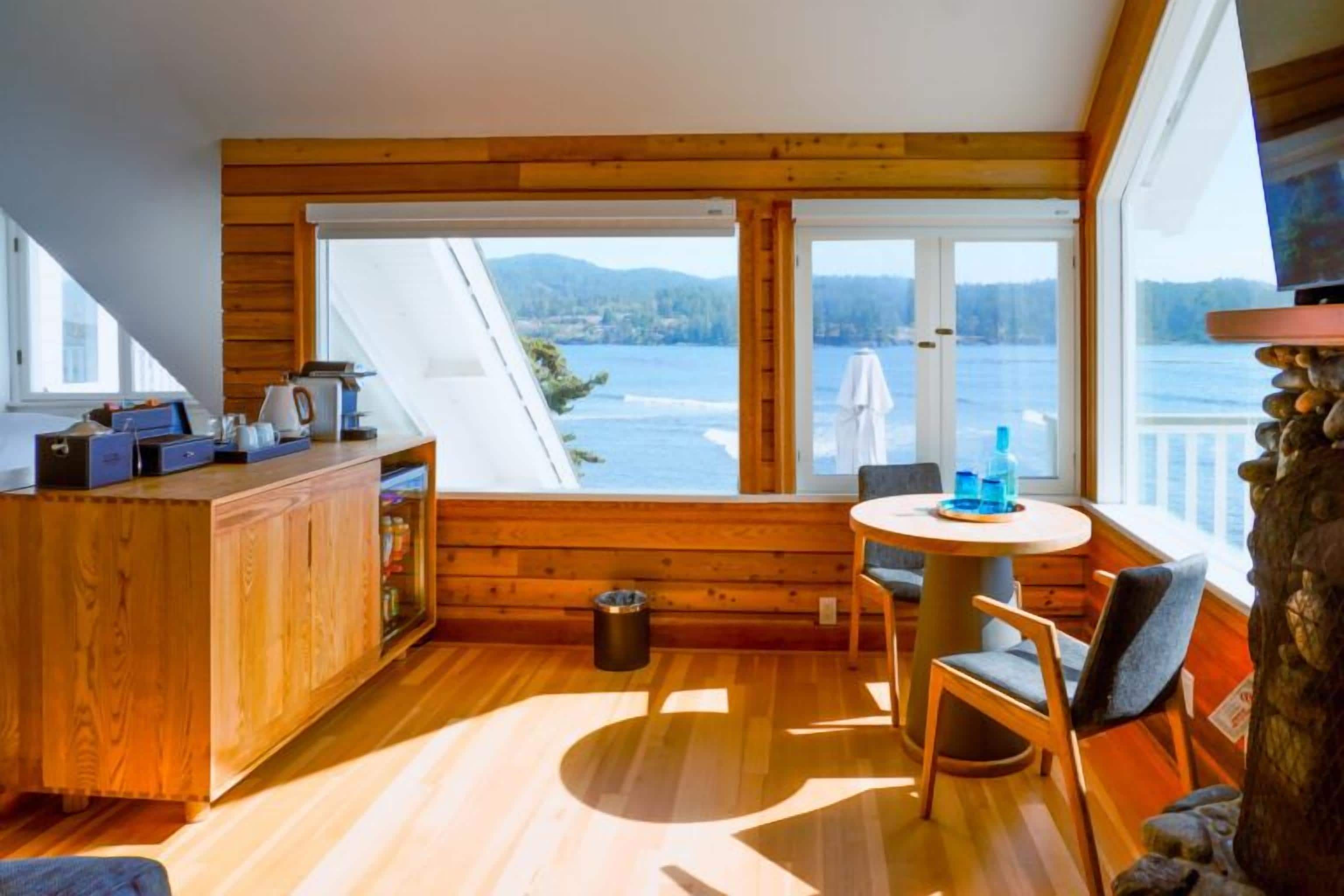 Uma cabana com interior de madeira, área de jantar com mesa e cadeiras, e vista para um lago através de amplas janelas.
