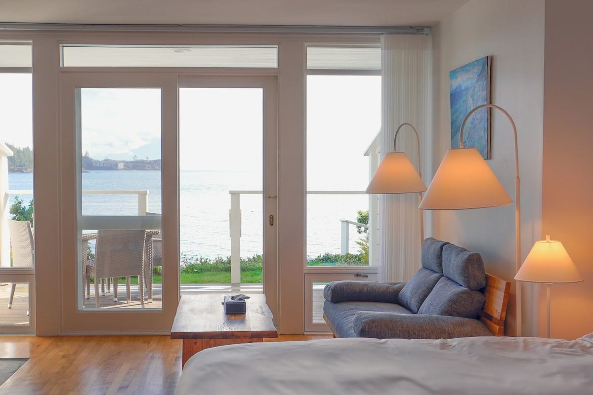 A bedroom with a large bed, a sofa, two lamps, and a view of the sea through a sliding door.