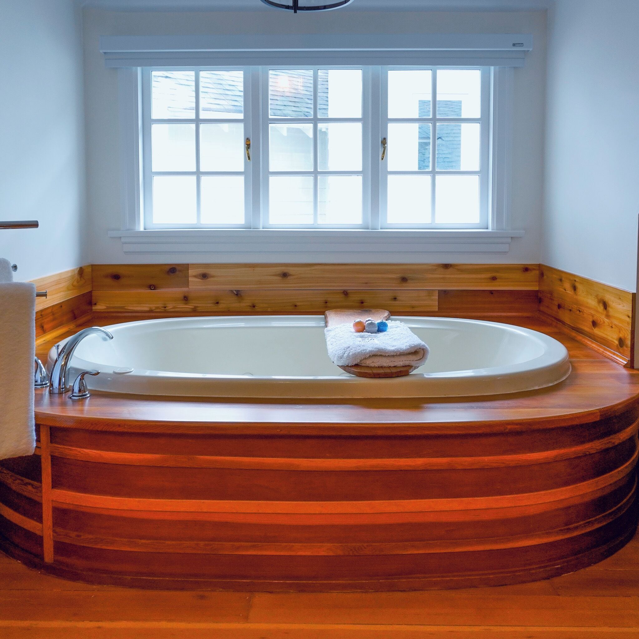 A large, oval-shaped bathtub with a wooden base, positioned under a window with multiple panes.