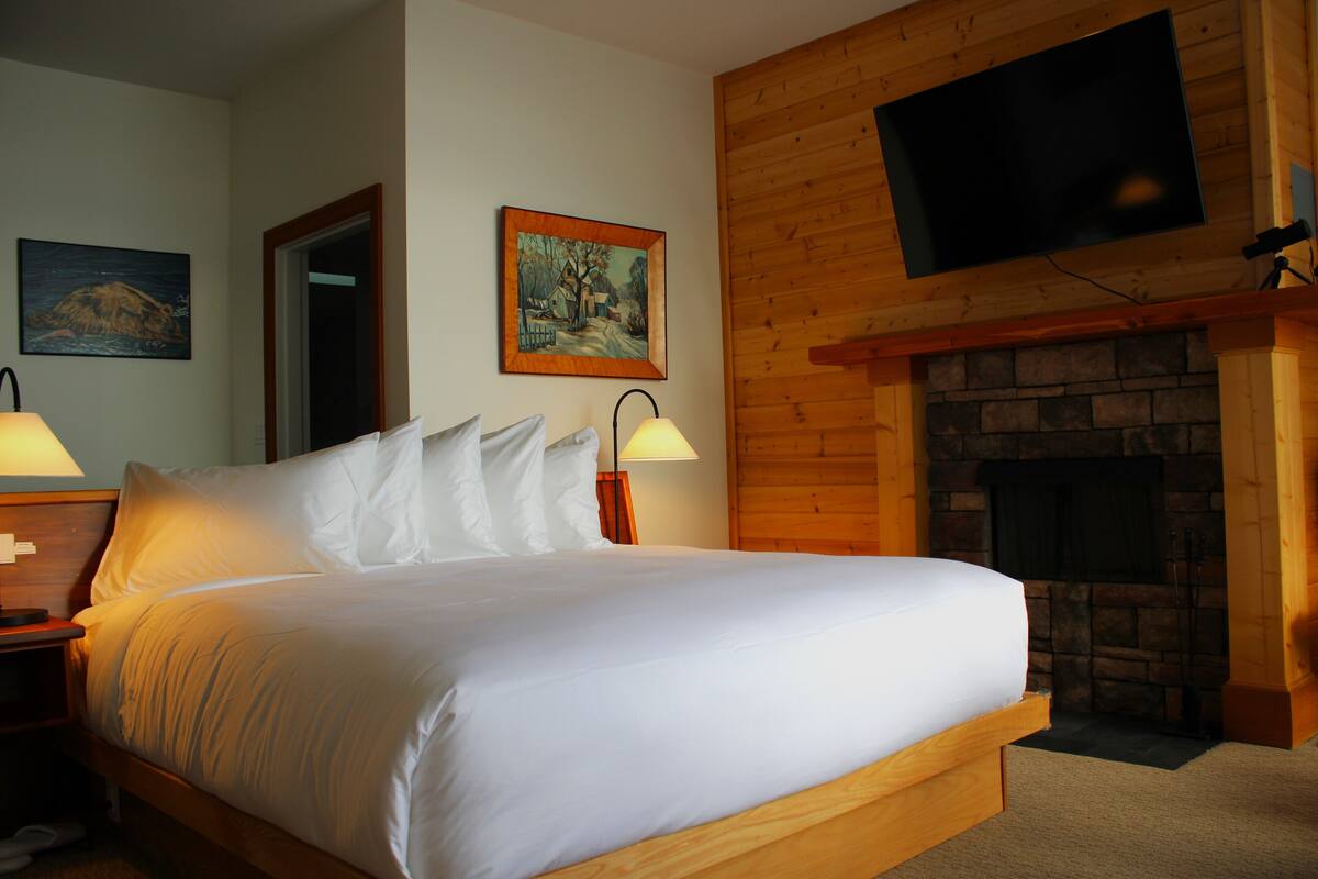 A bedroom with a large bed, a wooden headboard, a fireplace, a flat-screen TV mounted on the wall, and two bedside lamps.