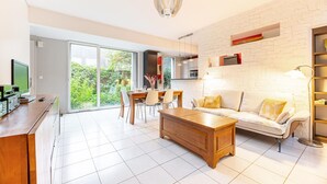 A modern living room with a white sofa, wooden coffee table, and a dining area with chairs and a table. There is a kitchen area with a microwave and cabinets, and a large window offering a view of greenery.