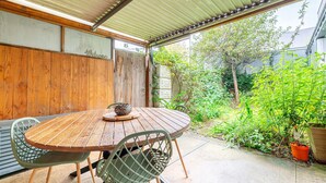 A wooden table with chairs under a metal roof, surrounded by greenery and a wooden fence.