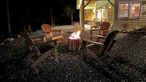 An outdoor seating area with four Adirondack chairs around a central fire pit, set on a gravel surface with a wooden building in the background. - Charming Cottage at Pretty Pond (Mount Desert)