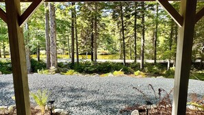 A covered outdoor area with a gravel path, surrounded by tall trees and a body of water. - Charming Cottage at Pretty Pond (Mount Desert)
