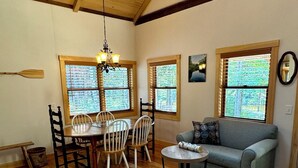 A spacious living room with wooden flooring, a dining table with chairs, a gray sofa, and large windows with blinds. - Charming Cottage at Pretty Pond (Mount Desert)