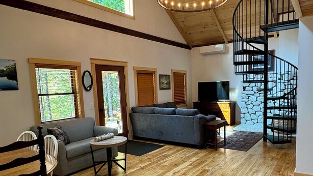 A spacious living room with wooden floors, a large chandelier, a spiral staircase, and large windows. - Charming Cottage at Pretty Pond (Mount Desert)