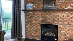 A living room with a brick fireplace, a framed picture above it, and a view through a large window. - On Hill Mountain Retreat (Kimberley)