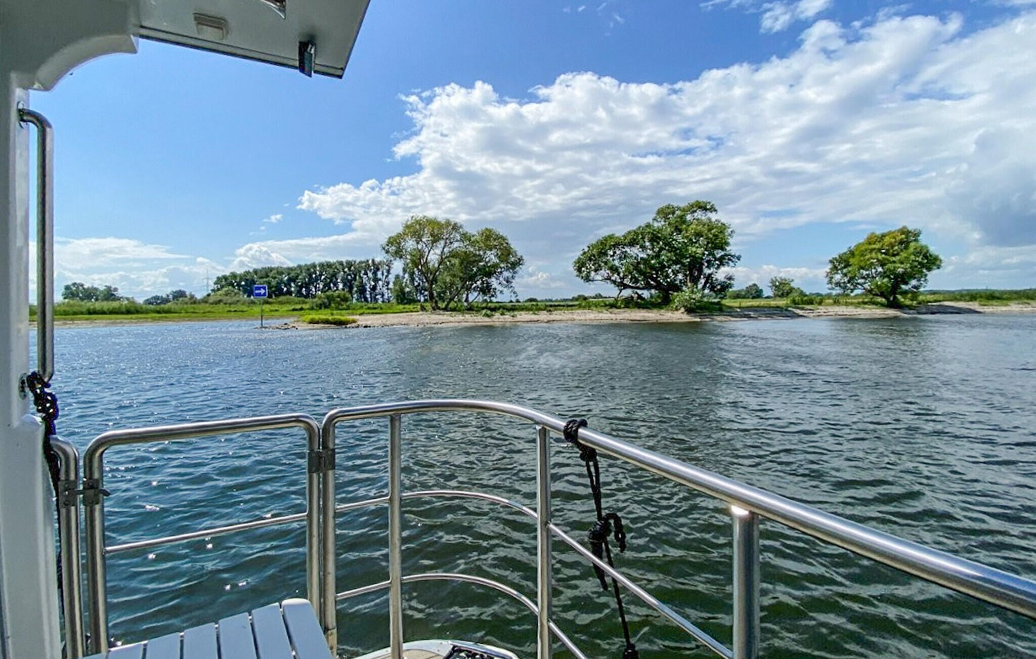 A boat's deck with metal railings and a view of a riverbank with trees and a sign.