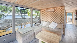Un intérieur chaleureux avec un coin salon confortable doté de coussins, de grandes fenêtres donnant sur une plage et un plafond en bois.