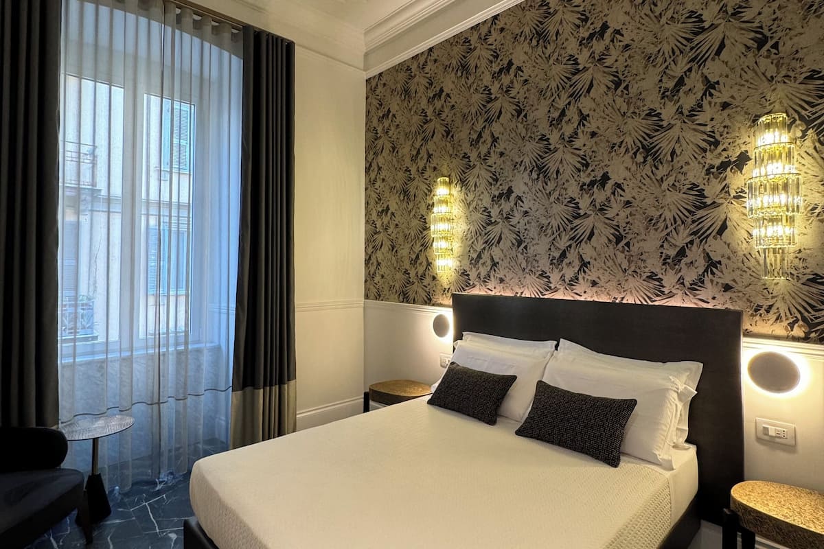 Habitación de hotel con una cama grande, dos lámparas de noche, una mesita pequeña y vistas a través de cortinas de gasa.