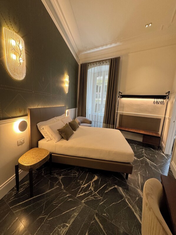 A modern hotel room with a large bed, a bedside table, a built-in bench with hooks, and a view of the outdoors through the window. - Lovely rooms (La Spezia)