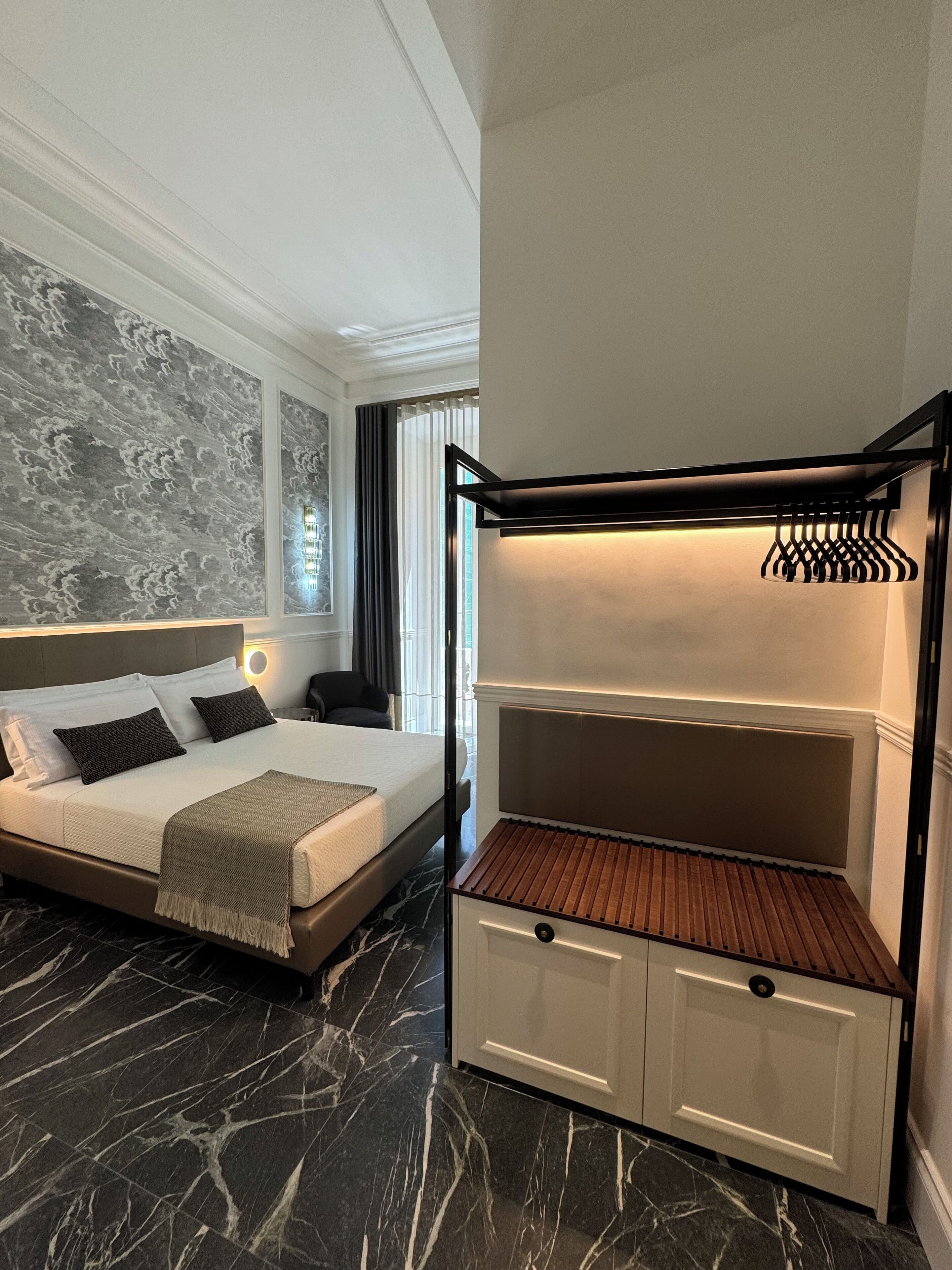 Una habitación de hotel con cama, un banco empotrado con espacio de almacenamiento, una lámpara de pared y suelo de mármol.