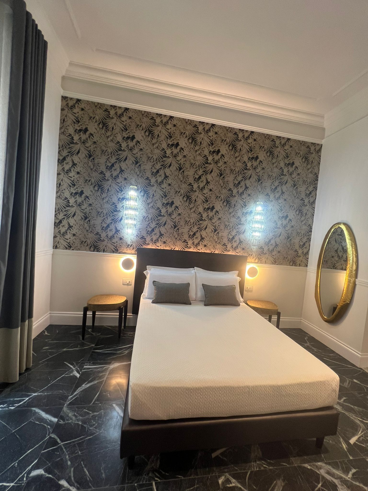 Een hotelkamer met een groot bed, twee nachtkastjes, een ronde spiegel en behang met een patroon.