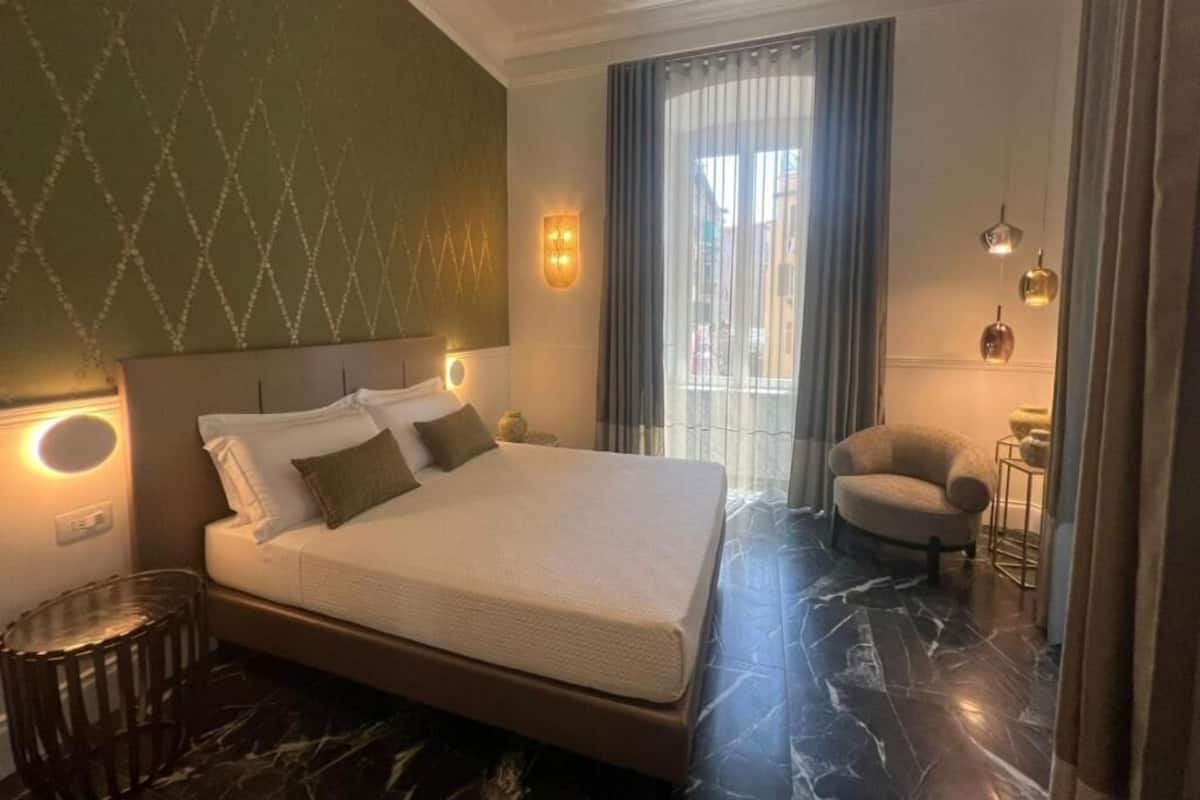Una habitación de hotel con una cama grande, un sillón pequeño, una mesita auxiliar y una ventana con cortinas.