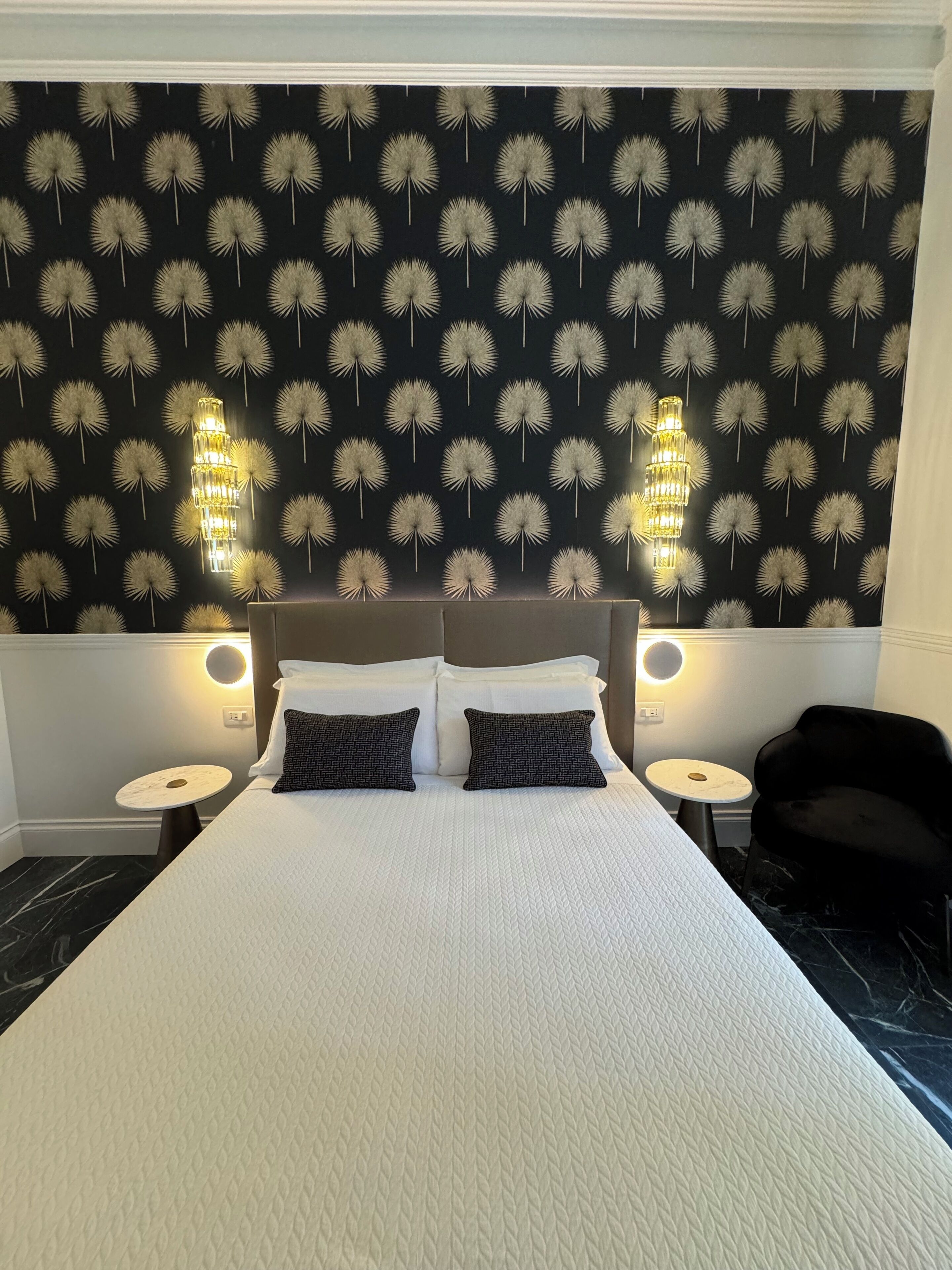 Habitación de hotel con una cama grande, dos mesitas de noche, un sillón negro y una pared con estampado de diente de león.