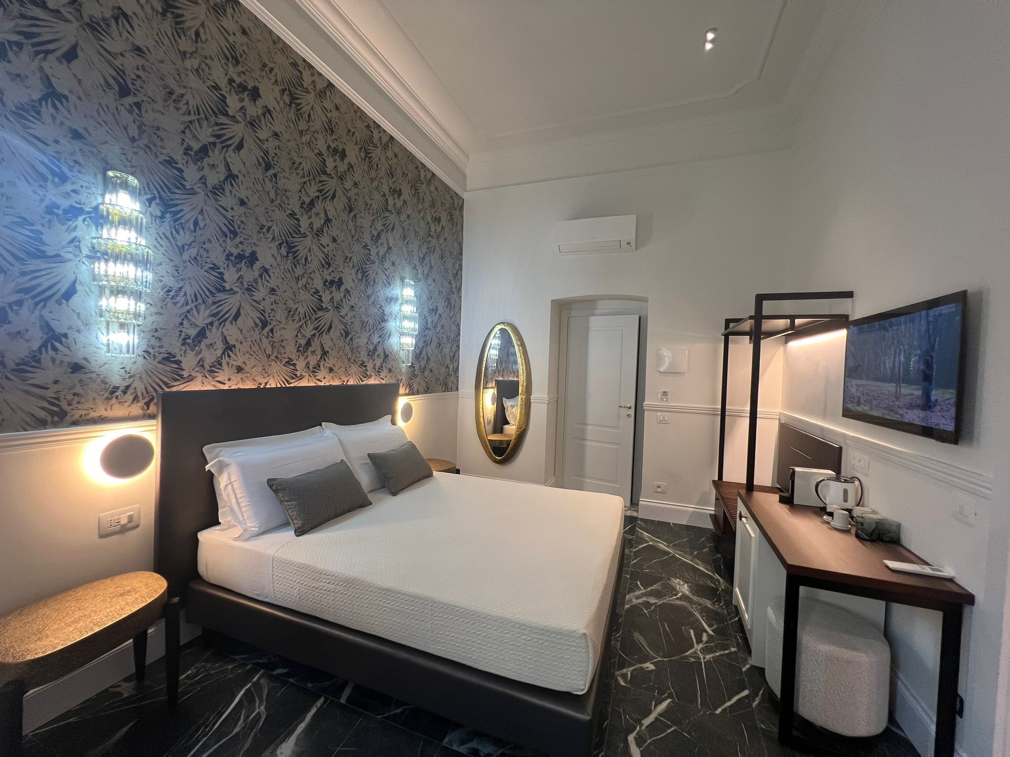 Una habitación de hotel moderna con una cama grande, una mesita de noche, un espejo, un televisor y un pequeño rincón de descanso.