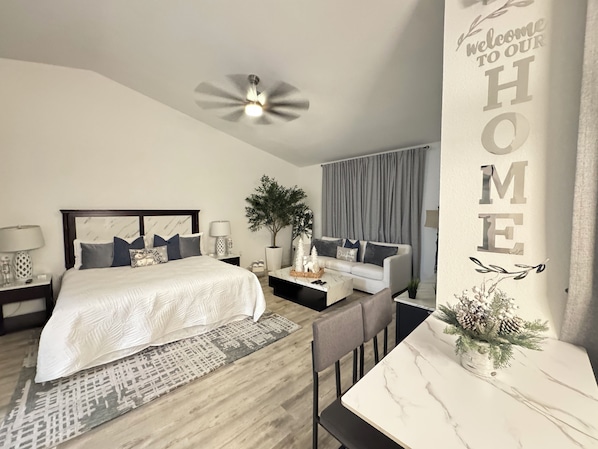A modern bedroom with a bed, bedside tables, a sofa, a dining table, a ceiling fan, and a wall with a 'Welcome to Our Home' sign.